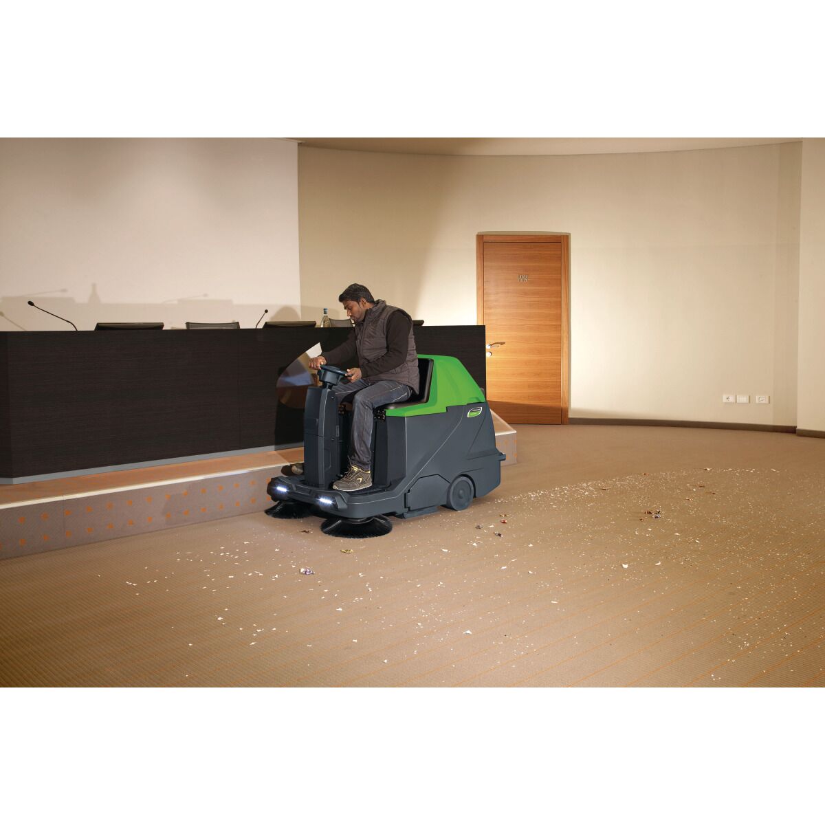 Person fährt die grün-schwarze Aufsitz-Kehrmaschine in einem Konferenzraum.