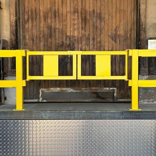 Gelbes Schutzgeländer mit Doppelflügeltor auf Rampe, montiert vor altem Holztor.