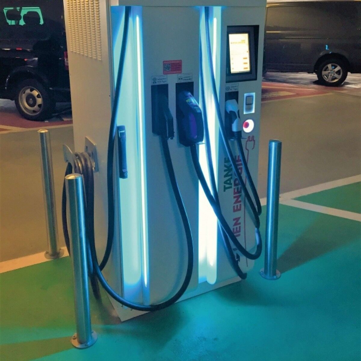 E-Ladestation mit Display und zwei Ladekabeln; drei Edelstahl-Poller als Rammschutz auf markierter Stellfläche, Fahrzeuge im Hintergrund.