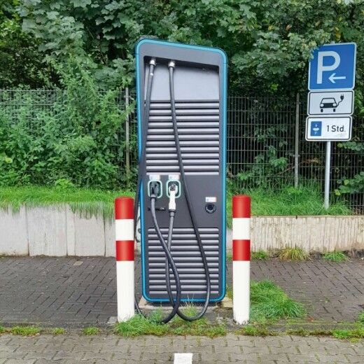 Zwei rot-weiße Rammschutz-Poller sichern eine E-Ladesäule an einem öffentlichen Parkplatz mit Hinweisschild.