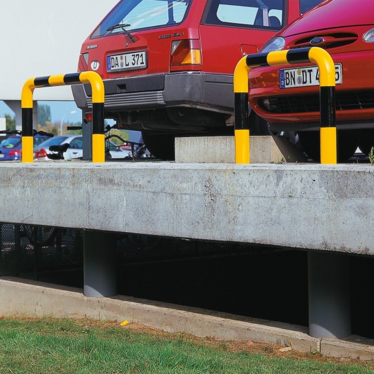 Foto: Zwei gelb-schwarz markierte Rammschutzbügel an einer Betonrampe vor parkenden roten Autos, Außenbereich.