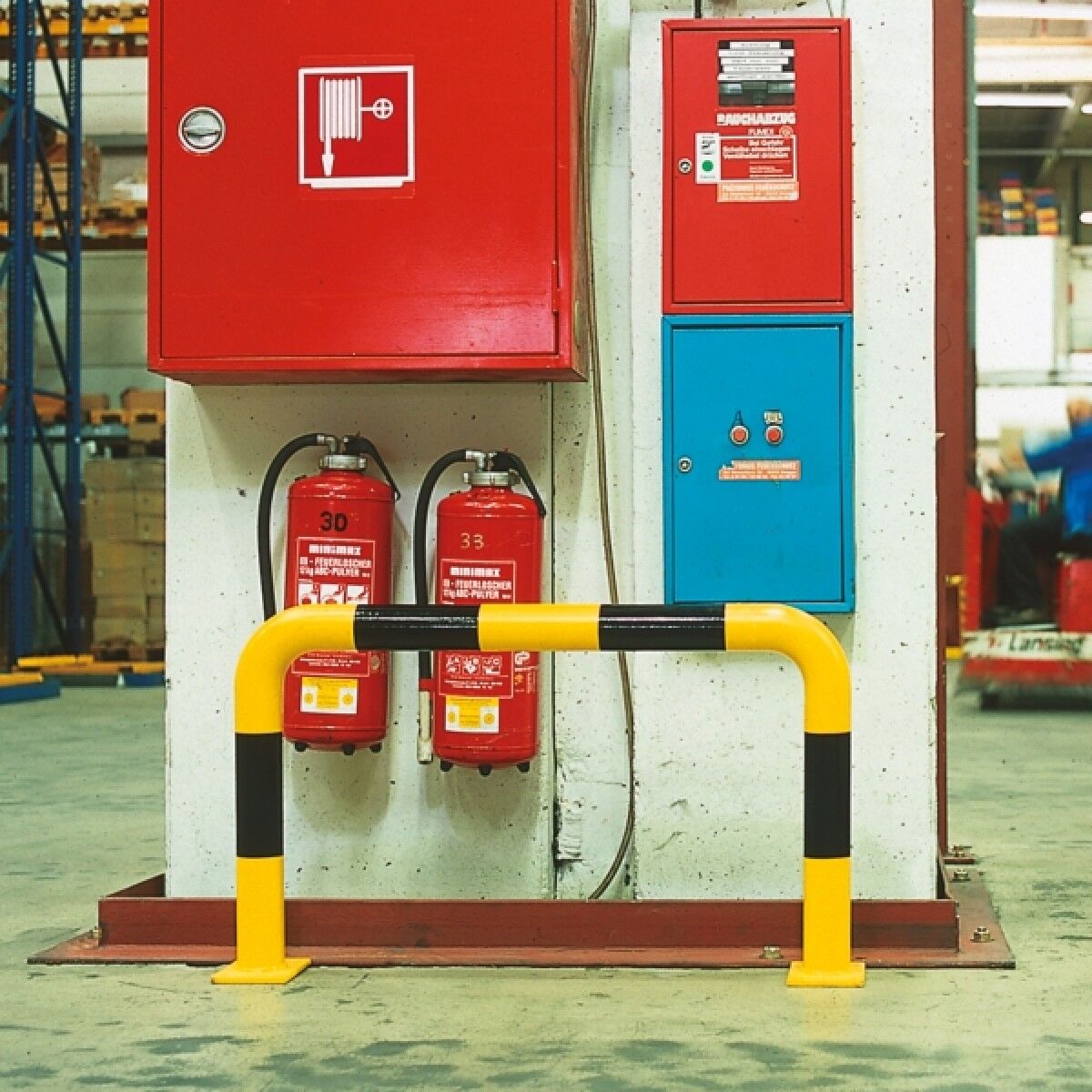 Gelb-schwarz gestreifter Rammschutzbügel vor Betonpfeiler; dahinter rote Wandkästen und zwei Feuerlöscher in Werkhalle.