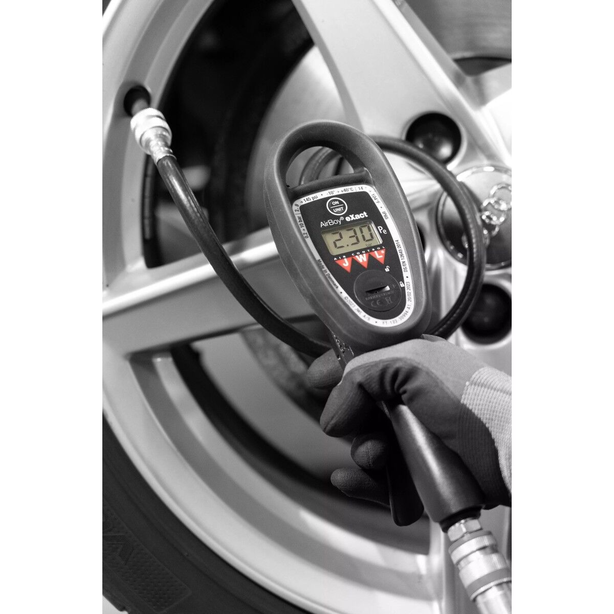 Schwarzweißfoto: Hand im Handschuh hält digitales Reifenfüllgerät vor Autofelge; Schlauch ist am Ventil angeschlossen.