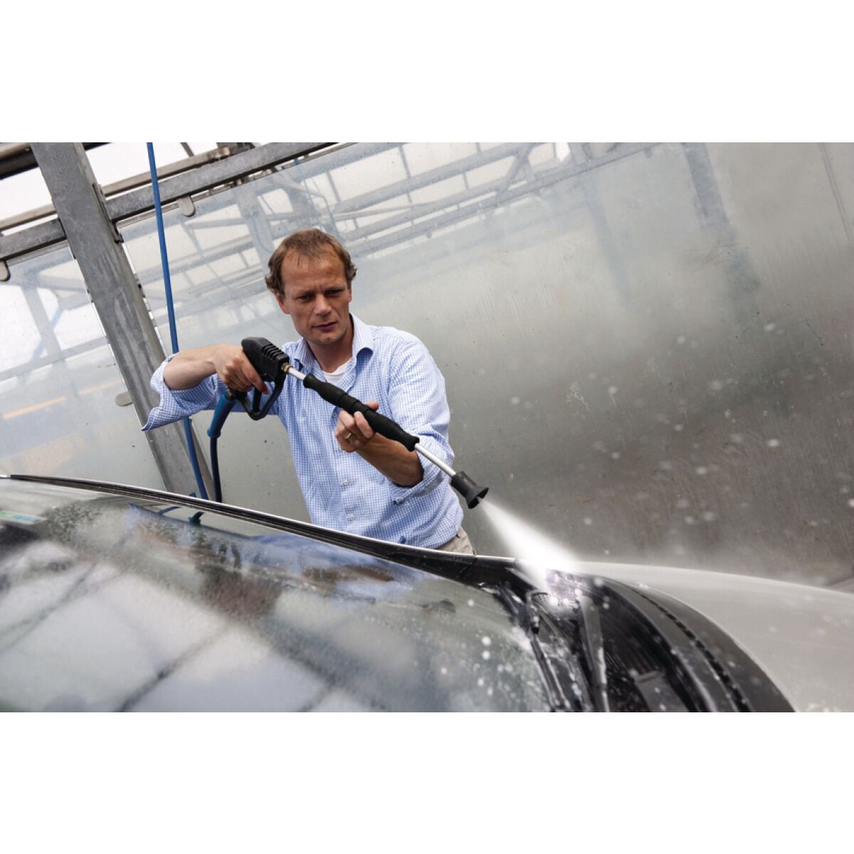 Person wäscht ein Auto mit Hochdruckreiniger in einer Waschanlage, kräftiger Wasserstrahl auf das Fahrzeugdach gerichtet.