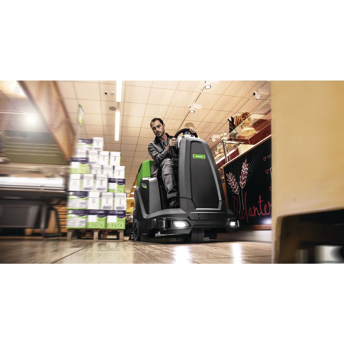 Eine Person steuert eine Aufsitz-Scheuersaugmaschine durch die Gänge eines Supermarkts mit gestapelten Waren.