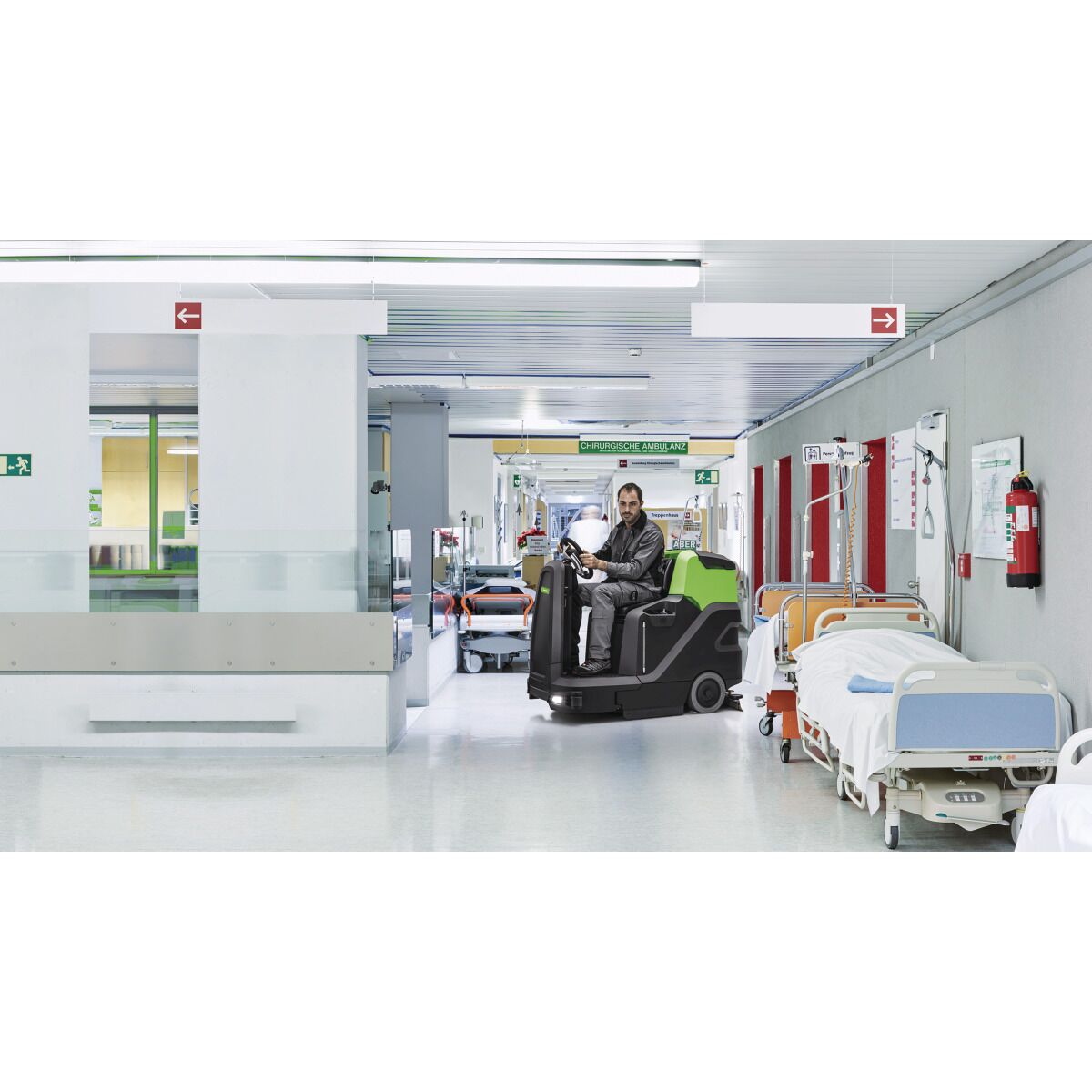 Eine Aufsitz-Scheuersaugmaschine fährt durch einen Krankenhausflur mit Betten und medizinischer Ausrüstung.