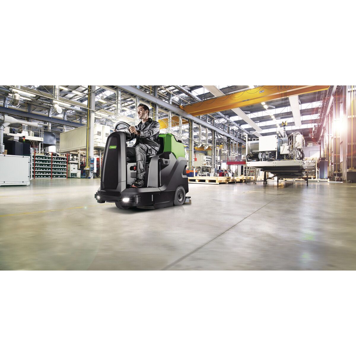 Eine Aufsitz-Scheuersaugmaschine wird in einer hellen Industriehalle mit Produktionsanlagen betrieben.