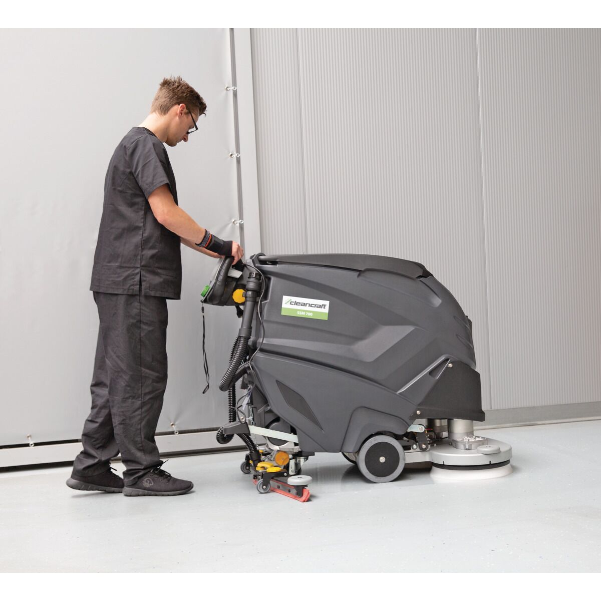 Person bedient graue Bodenreinigungsmaschine in einer Halle mit hellem Boden und grauer Wand.