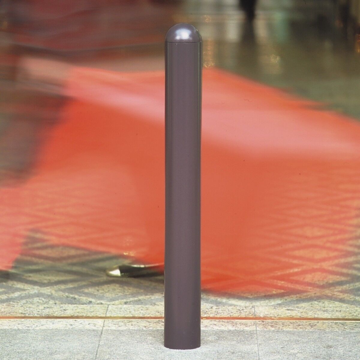 Grauer Poller mit halbkugeligem Kopf auf Steinboden; Hintergrund rot und unscharf.