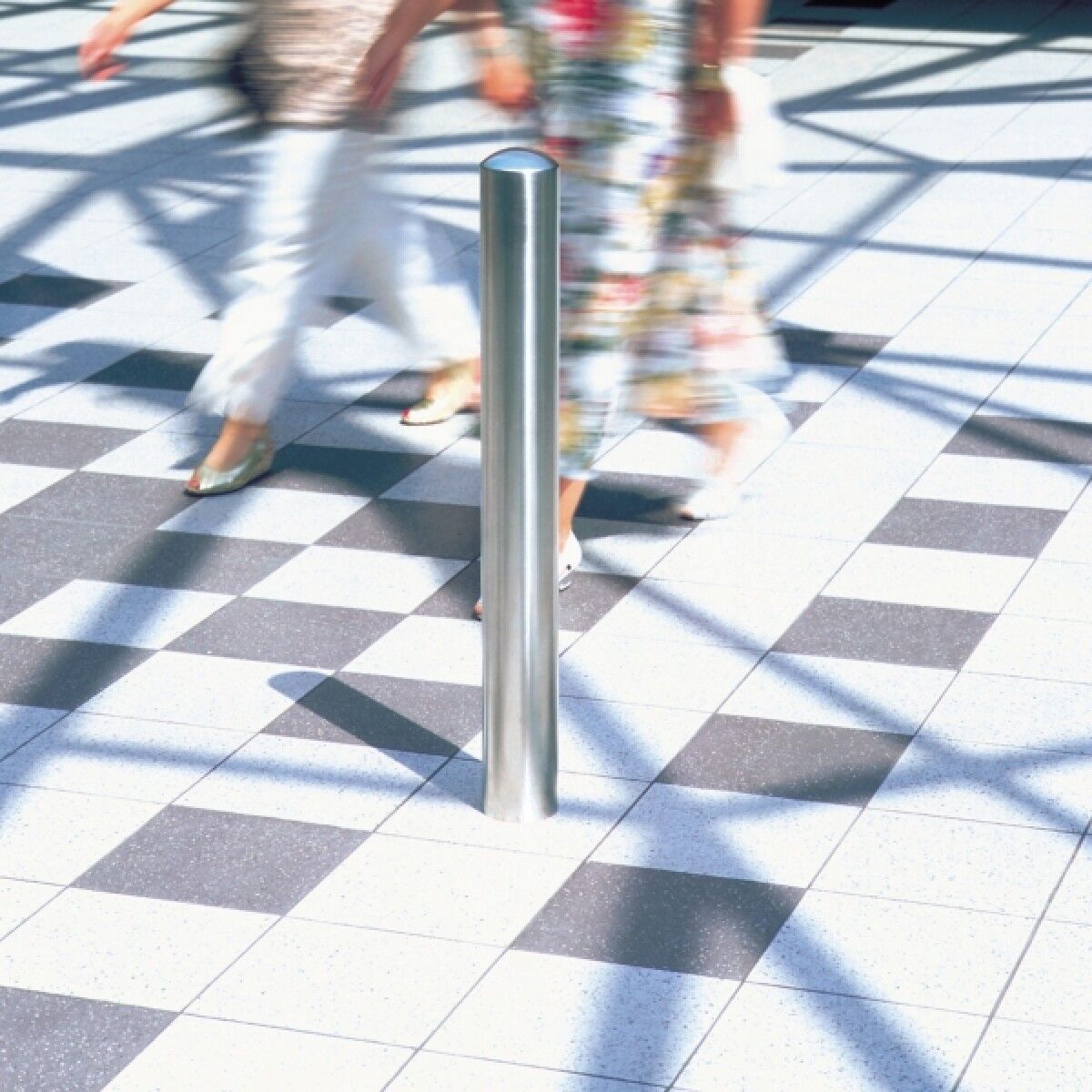 Edelstahl-Poller in gefliestem Innenraum; unscharfe Personen gehen vorbei, Schattengitter auf dem Boden sichtbar.