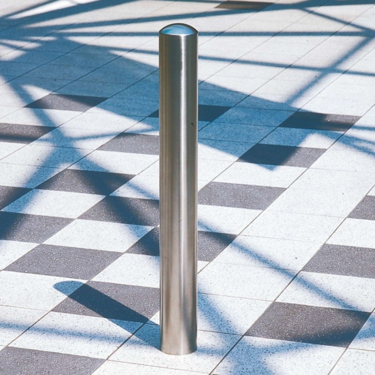 Edelstahl-Poller als runder Pfosten auf grau-weiß gefliestem Boden, mit schrägen Schattenlinien im Hintergrund.