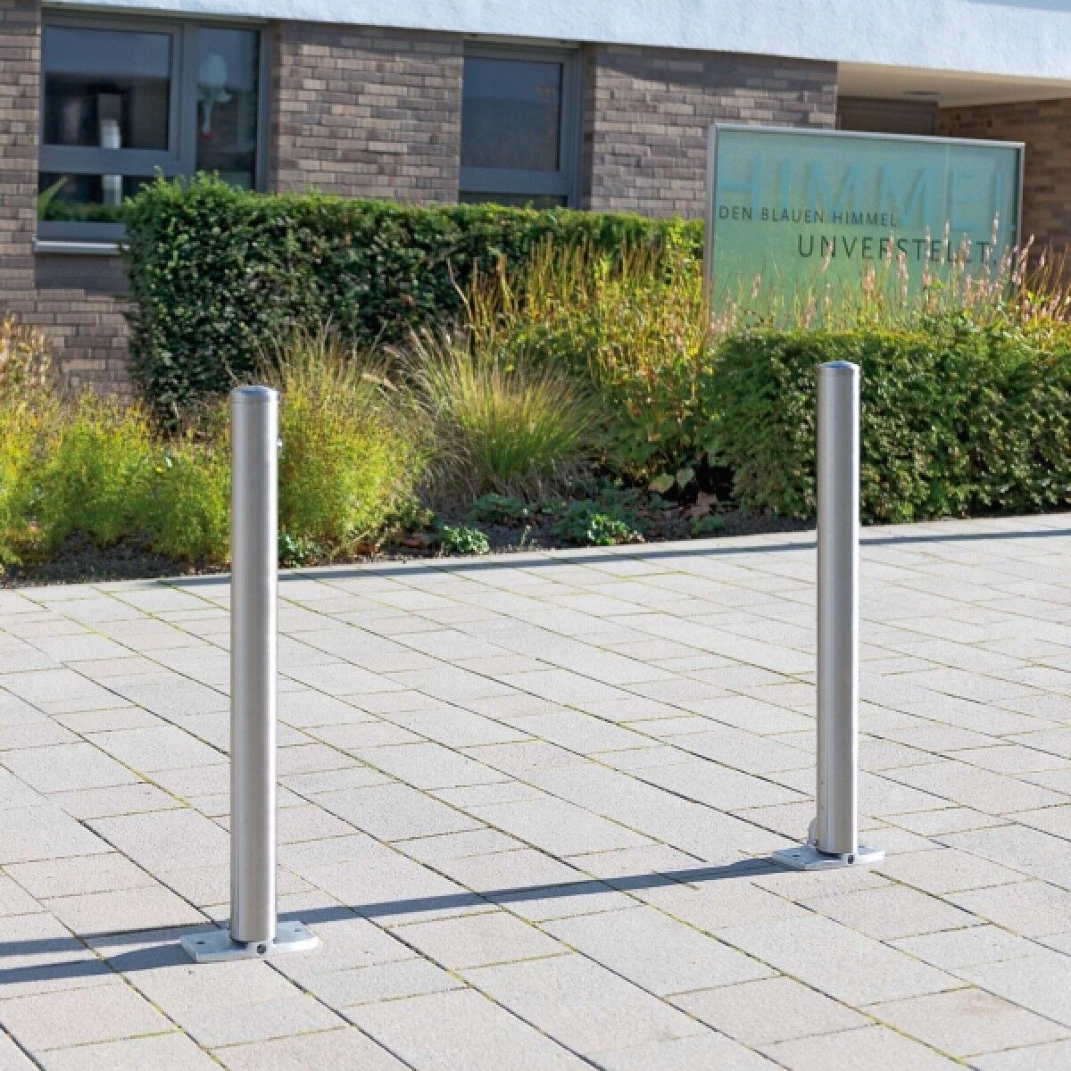 Zwei Edelstahlpfosten auf Sockelplatten im Pflaster, vor Hecken und Gebäudefassade mit Schild.
