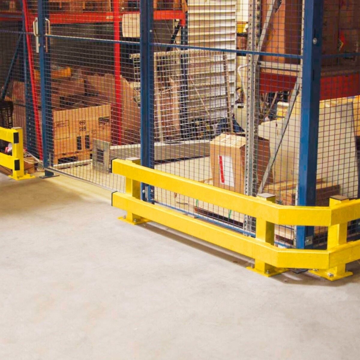 Gelber zweireihiger Rammschutz entlang eines Drahtgitter-Zauns, an einer Ecke montiert; Kartons hinter dem Zaun.