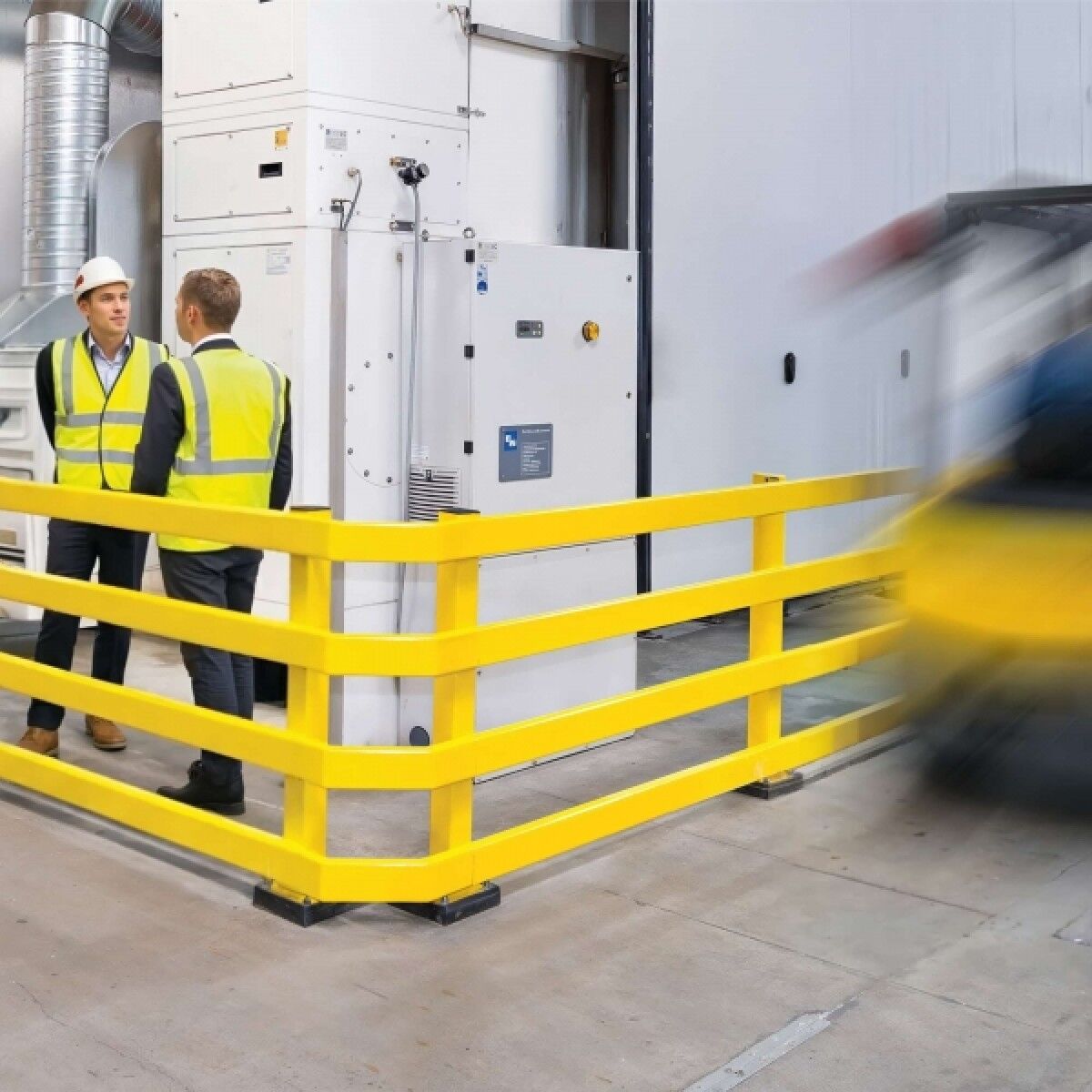 Gelbes vierreihiges Rammschutzgeländer um weiße Industrieanlage; zwei Männer in Warnwesten sprechen, rechts fährt ein unscharfer Gabelstapler vorbei.