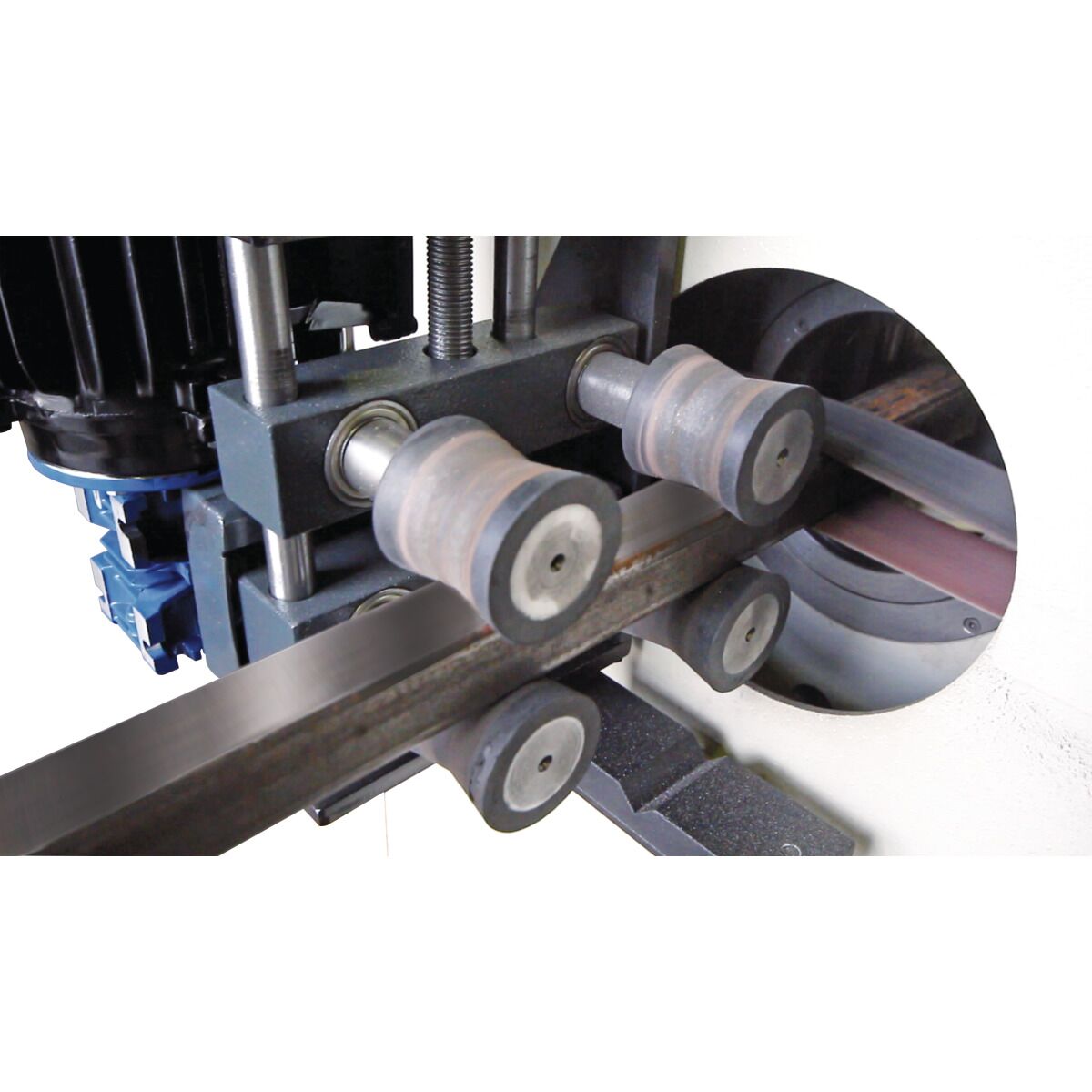 Detailansicht der Vorschubrollen einer Metallbandschleifmaschine beim Schleifen eines rechteckigen Metallprofils.