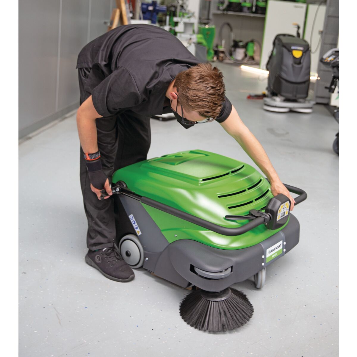 Person mit schwarzer Arbeitskleidung bedient eine grün-schwarze Kehrmaschine in einer Werkhalle.