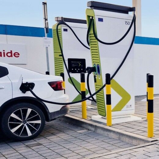 Weißes Elektroauto wird an zwei Ladesäulen mit schwarzen Ladekabeln auf einem gepflasterten Parkplatz geladen.