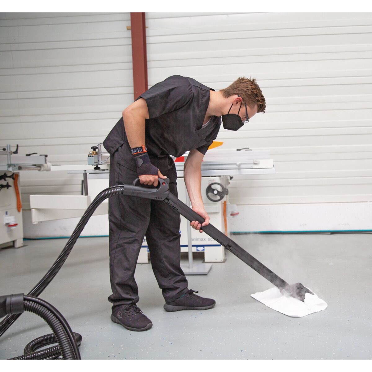 Person in Schutzkleidung und Maske reinigt mit Dampfreiniger und Bodendüse einen hellen Werkstattboden.