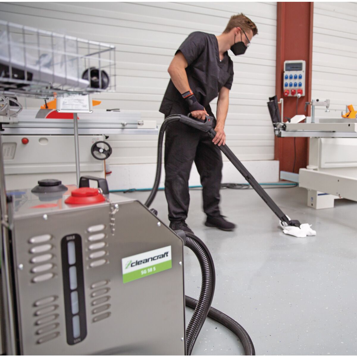 Person in schwarzer Arbeitskleidung nutzt einen Dampfreiniger auf Industrieboden, Gerät steht im Vordergrund.