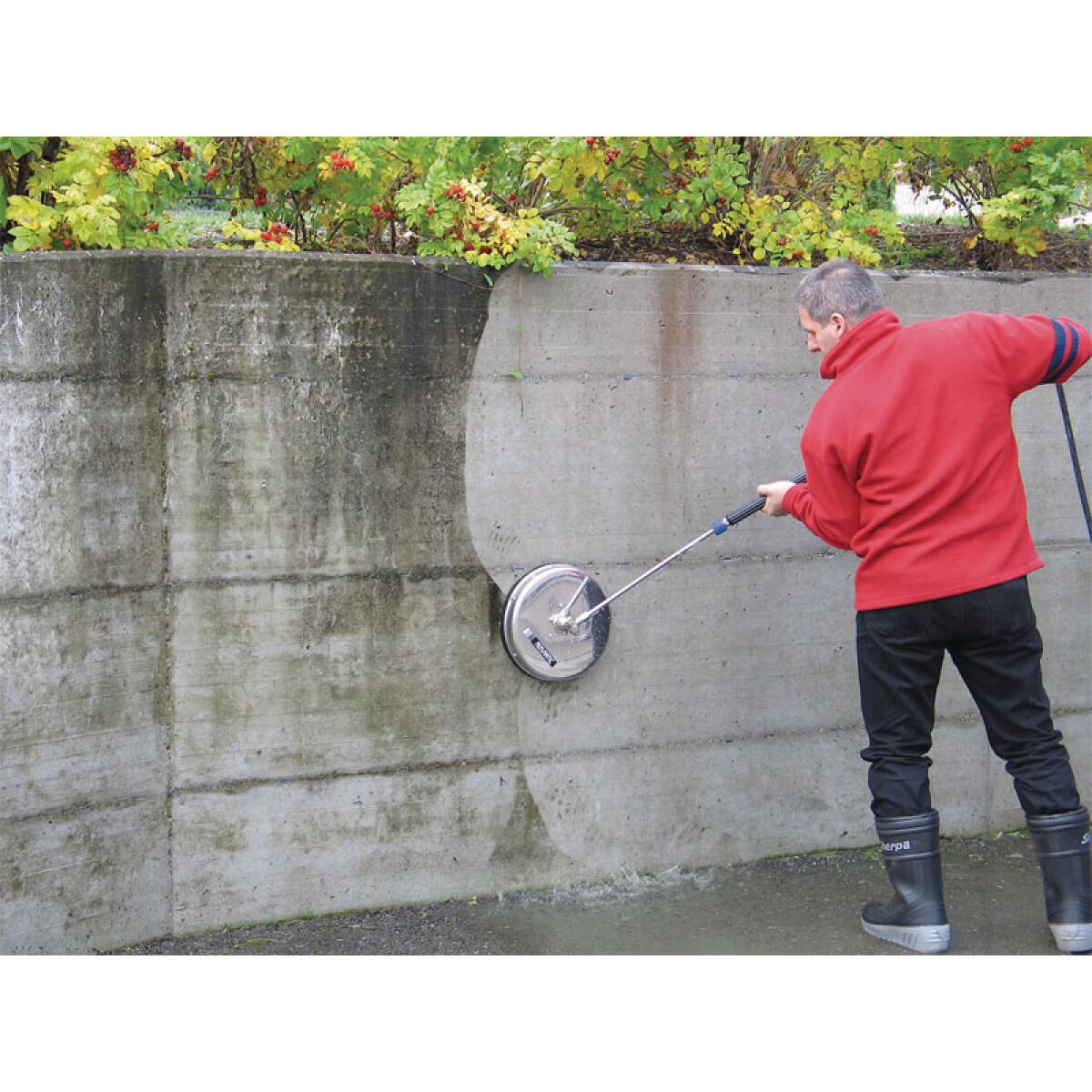 Person reinigt eine verschmutzte Betonwand mit einem Hochdruckreiniger und einer rotierenden Flächenreinigerdüse.